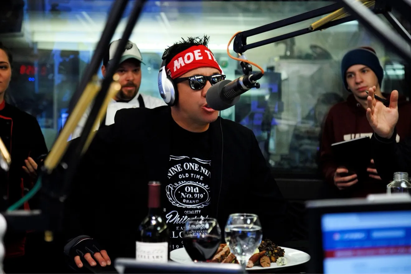 Moe Train on a broadcast mic in the studio, red MOE TRAIN headband and headphones on, a steak plate and wine glasses on the desk in front of him