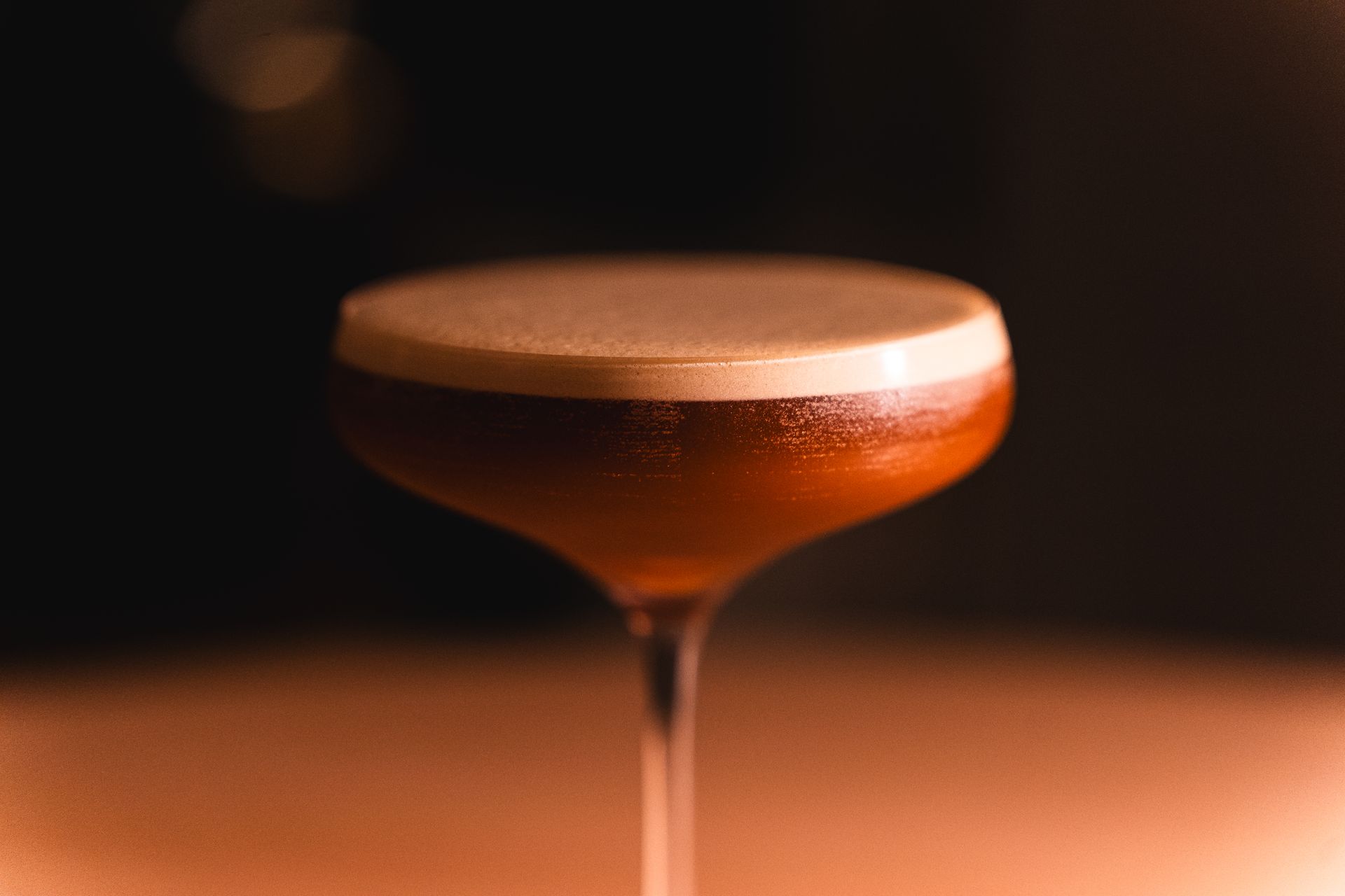 A cocktail coupe glowing amber against a dark bar