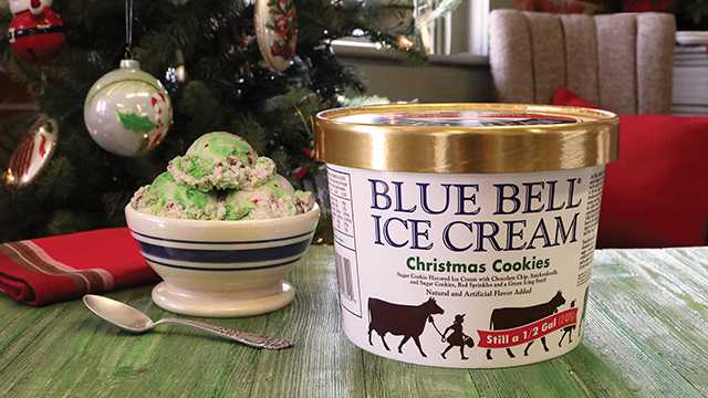 A half gallon carton of Blue Bell Christmas Cookies Ice Cream with its gold lid next to a bowl of scoops with a green icing swirl, in front of a decorated Christmas tree