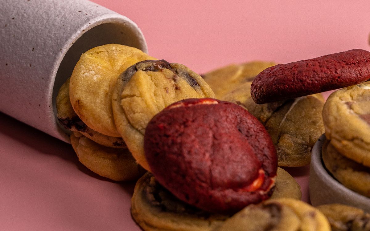 Assorted cookies, including deep red ones, spilling from a container on a pink background