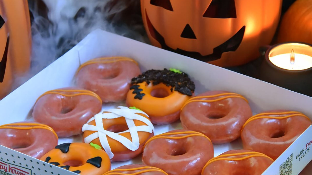 An open box of Krispy Kreme All Jacked Up doughnuts with orange glazed rings and three decorated Halloween doughnuts in front of a lit jack-o-lantern