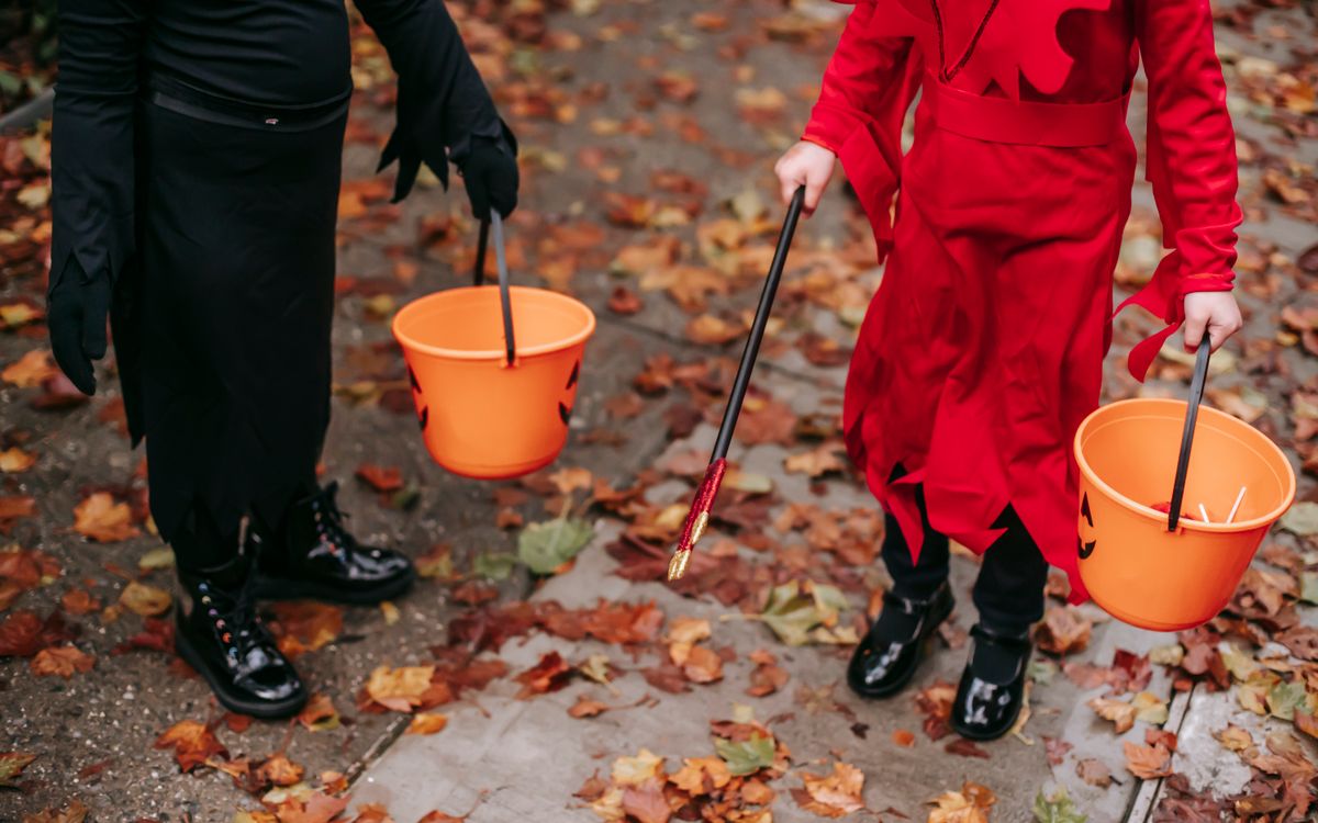 Two children in Halloween costumes holding orange pumpkin buckets on a leaf covered sidewalk