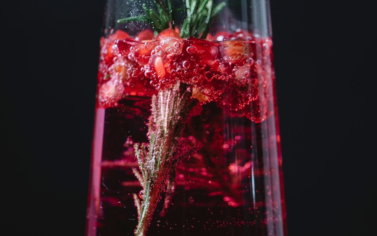 A glass of sparkling red pomegranate drink with pomegranate seeds and a rosemary sprig against a dark background