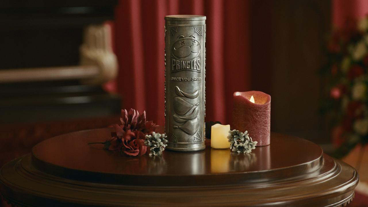 The metal Pringles urn from the Once You Drop contest on a wooden table with candles and flowers, engraved with the Pringles name and a stack of crisps