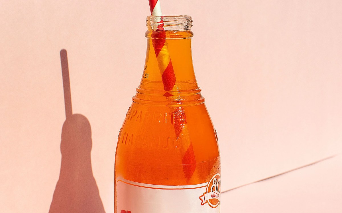 A glass bottle of orange soda with a striped straw against a pink background