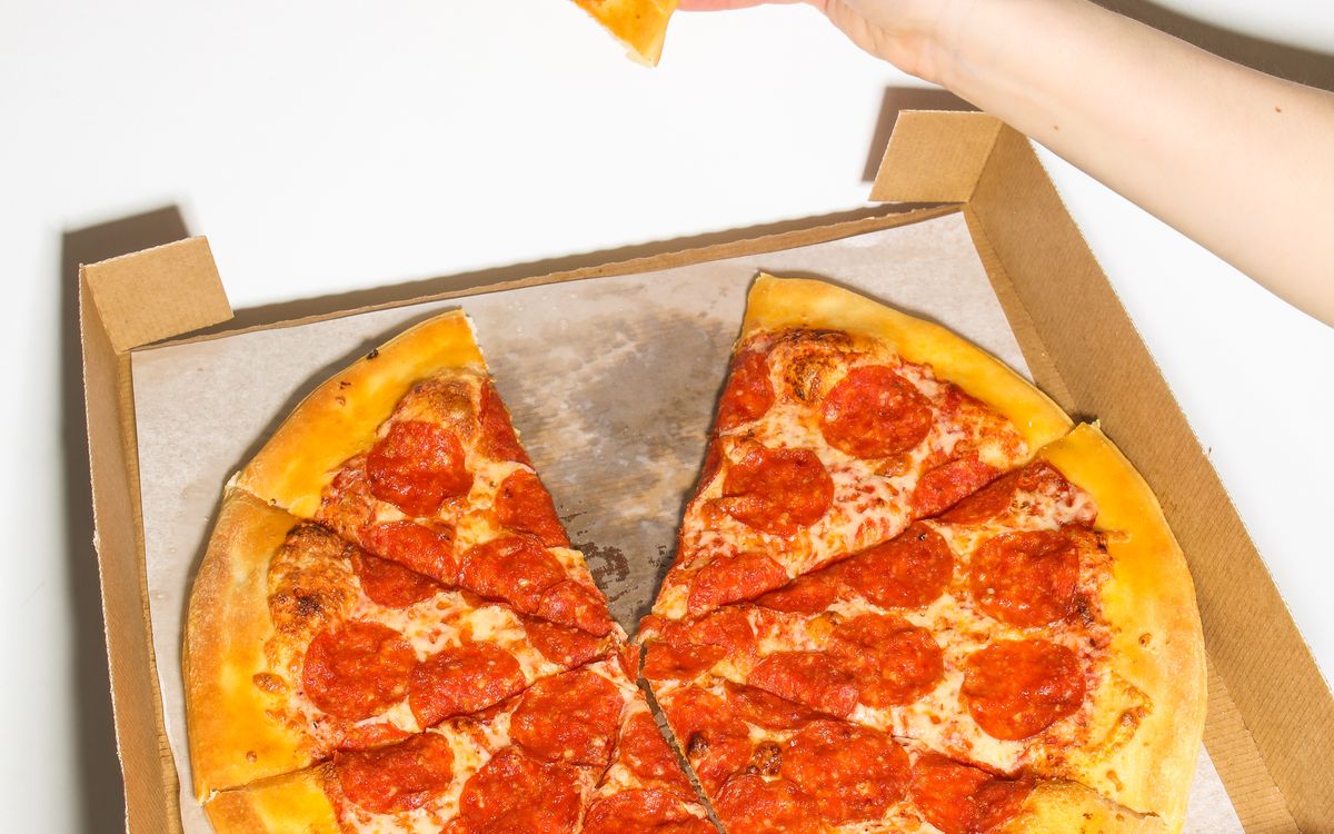 A pepperoni pizza in an open delivery box with one slice being lifted away