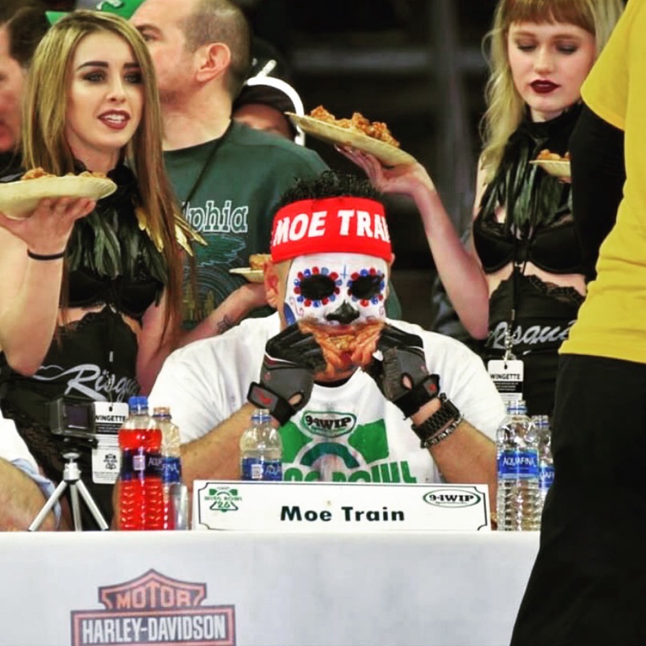 Moe Train biting into a wing at the Wing Bowl 26 table, his Moe Train nameplate in front of him and Wingettes serving plates behind