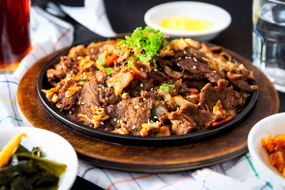A hot plate of beef bulgogi with sliced beef, sesame seeds, and green onions, with small bowls of side dishes around it