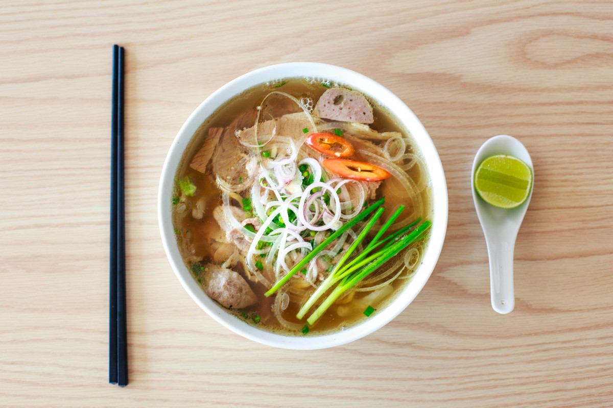 A bowl of pho from above with rice noodles, sliced meat, white onion, green onions, and red chili in clear broth, chopsticks beside the bowl and a lime wedge on a spoon