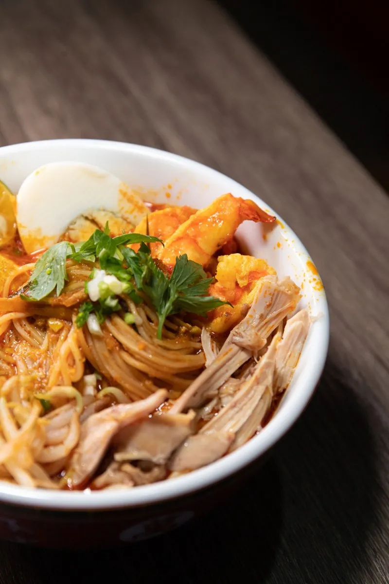 A bowl of curry laksa with shrimp, shredded chicken, egg, and noodles in orange coconut broth