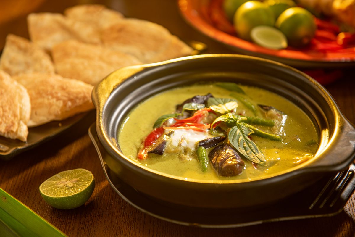 A bowl of Thai green curry with basil leaves, red chili, green beans, and eggplant in a green coconut sauce, with a lime half on the side