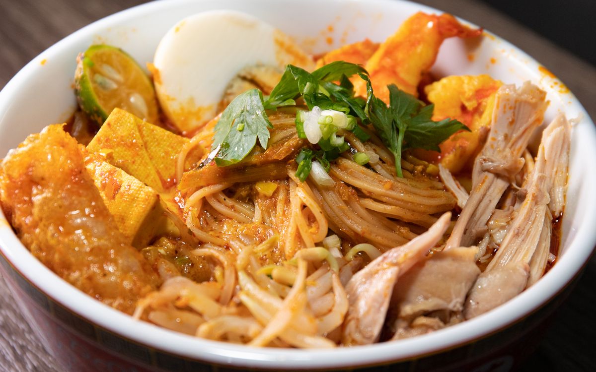 A bowl of rice noodles in a spicy orange gravy with egg, tofu, and herbs
