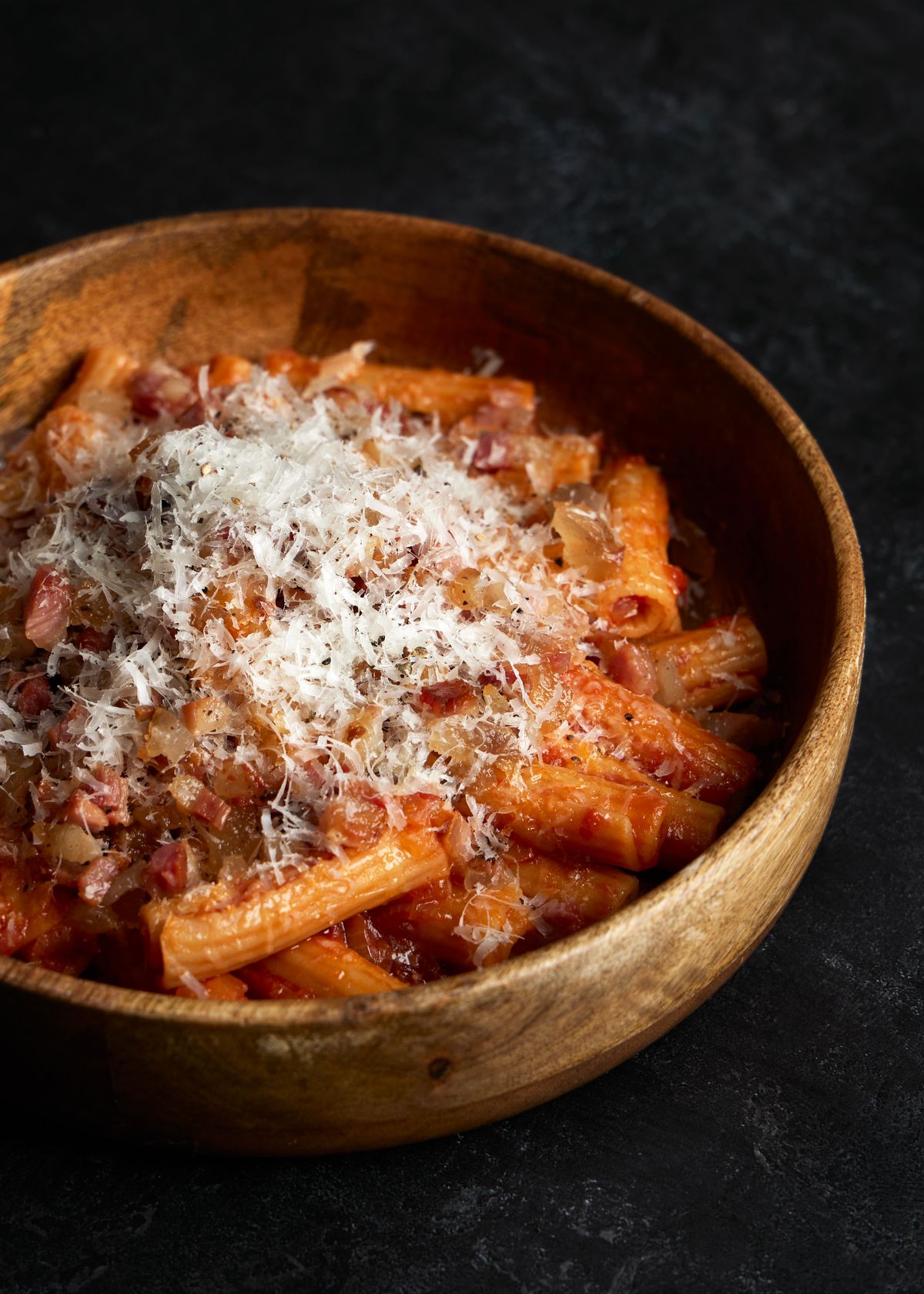 A wooden bowl of rigatoni in a creamy tomato sauce topped with grated Parmesan and crispy pork pieces