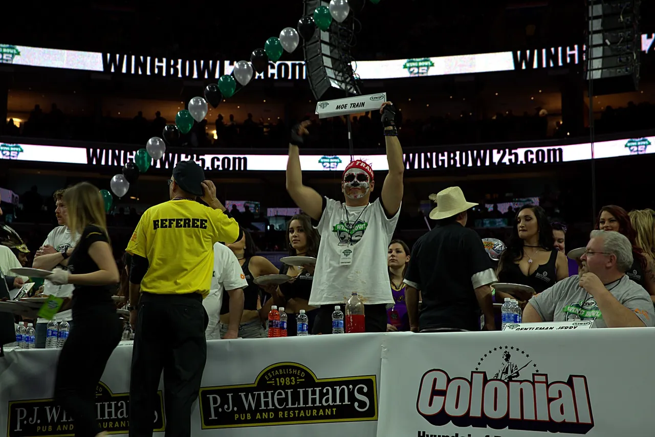 Moe Train at Wing Bowl 25 holding his MOE TRAIN name card overhead at the competition table, sugar-skull face paint and red MOE TRAIN headband, arena ring boards and a balloon arch behind him